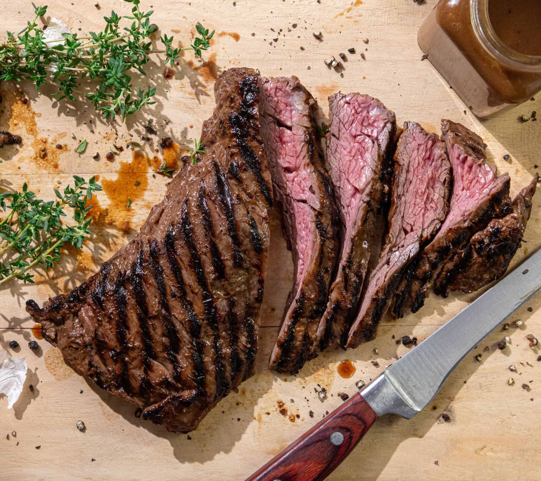 Sliced grilled steak on a wooden cutting board with fresh herbs, peppercorns, and a jar of sauce nearby.