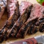 Sliced steak with a charred exterior and pink interior on a cutting board, knife placed beside it.