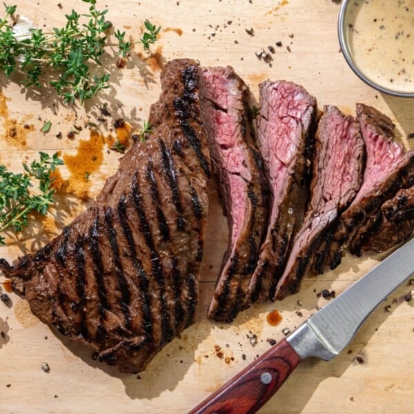 Grilled steak sliced on a wooden board with a knife, fresh herbs, black pepper, and a small bowl of sauce.