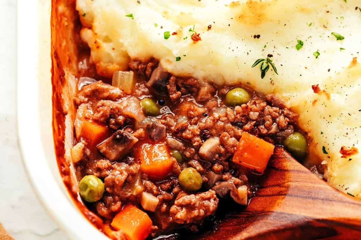 Ground meat, carrots, peas, and gravy topped with mashed potatoes in a baking dish; wooden spoon.