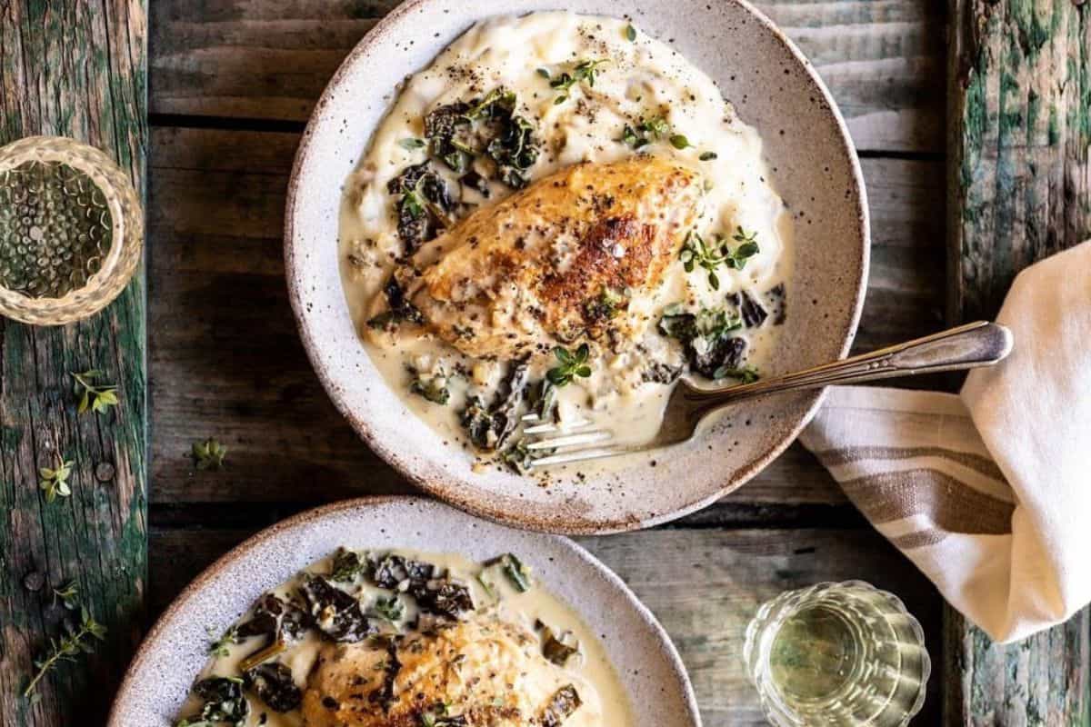 Two bowls of creamy chicken with herbs and mushrooms sit on a rustic wooden table, alongside two glasses of white wine and a striped napkin.