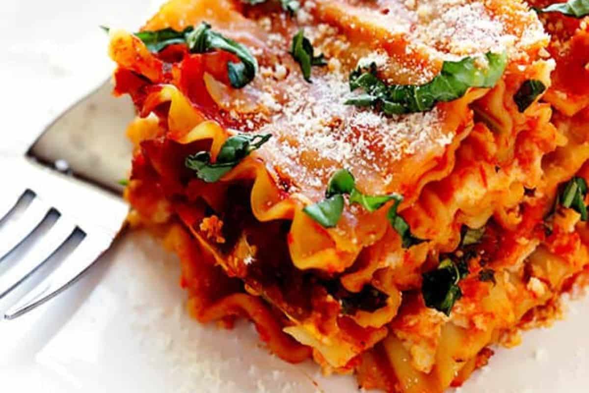 A slice of lasagna showing layers of pasta, tomato sauce, melted cheese, and fresh basil on top, placed on a white plate with a fork beside it.