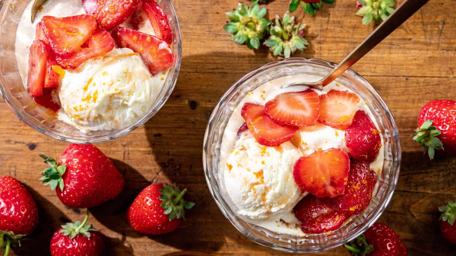 Two glass bowls of vanilla ice cream with sliced strawberries, on a wooden surface with whole strawberries and strawberry tops nearby.
