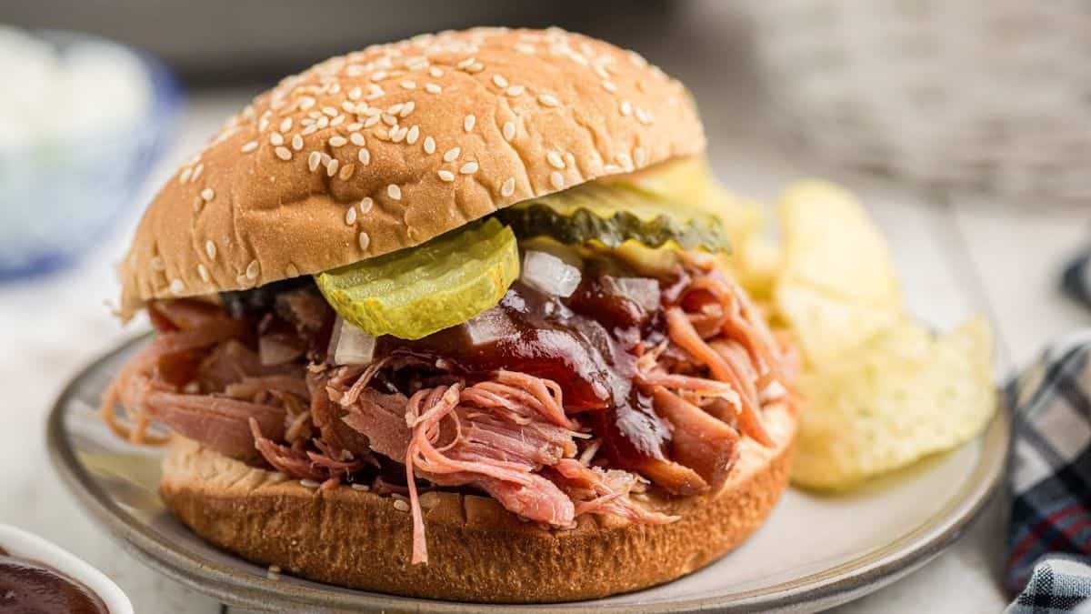 A pulled pork sandwich with barbecue sauce, sliced pickles, and onions on a sesame seed bun, served with potato chips on a plate.