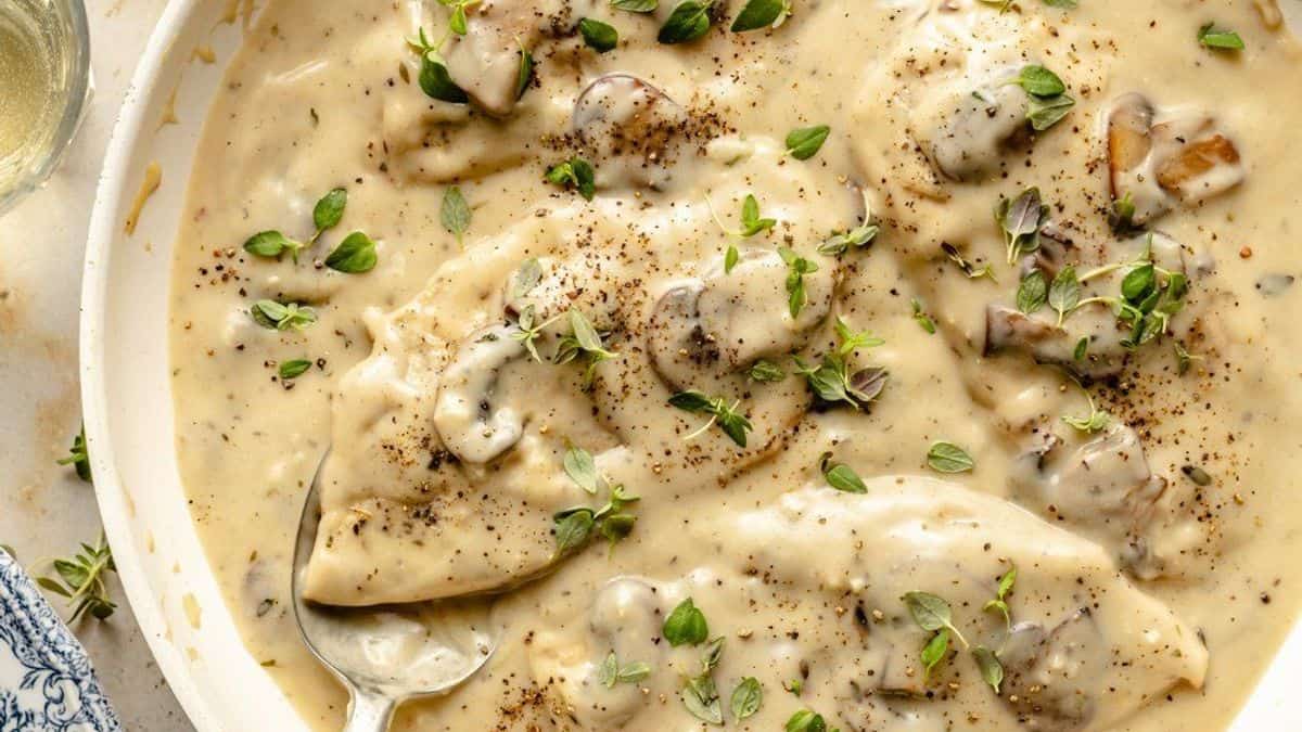 Chicken breasts covered in creamy mushroom sauce, topped with herbs and black pepper, on a white dish with a spoon.