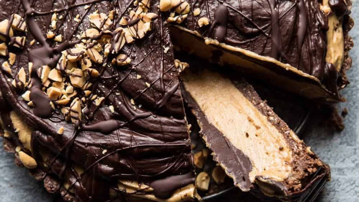 A chocolate peanut butter pie with a slice removed, revealing layers, topped with chopped peanuts and drizzled chocolate.