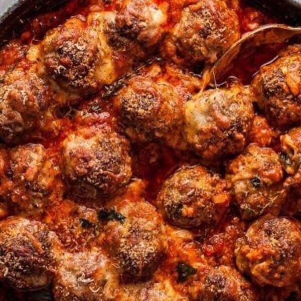 Baked meatballs in tomato sauce topped with melted cheese in a skillet, a spoon resting on the edge.