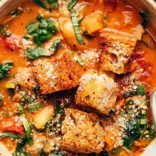 A bowl of vegetable soup with leafy greens, tomatoes, large croutons, grated cheese on top, and a spoon.
