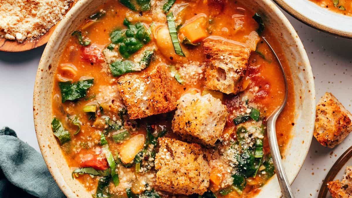 A bowl of vegetable soup with leafy greens, tomatoes, large croutons, grated cheese on top, and a spoon.