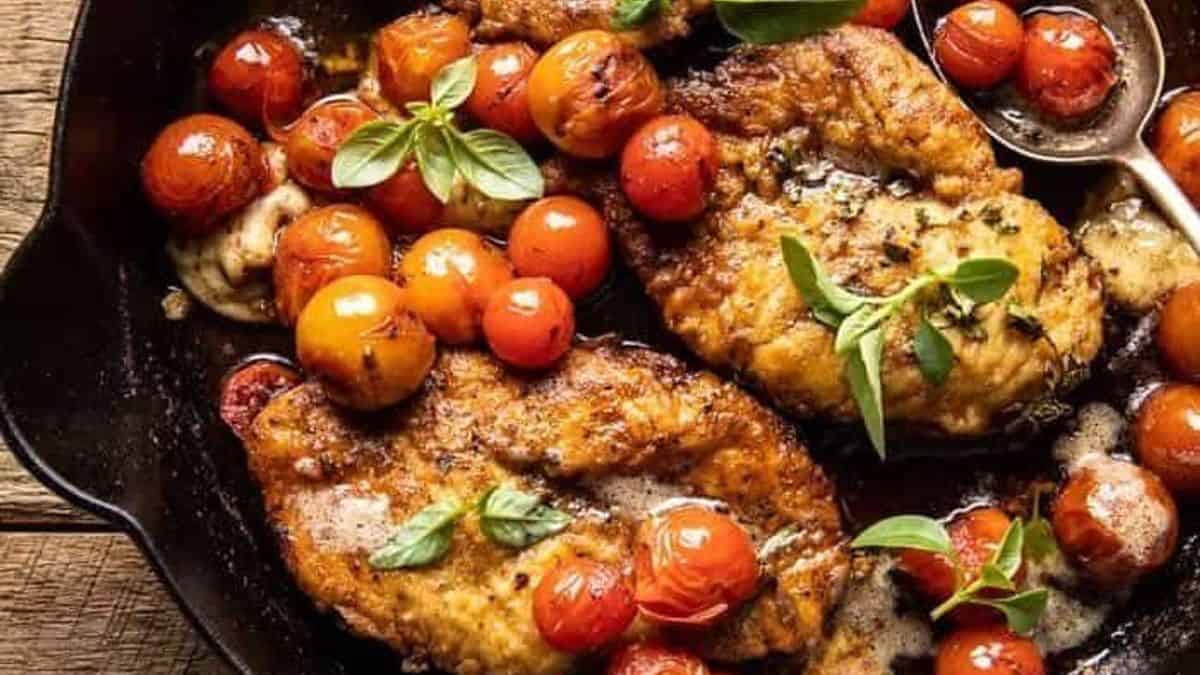 Pan-seared chicken breasts, roasted cherry tomatoes, fresh basil, and light sauce in a black skillet.