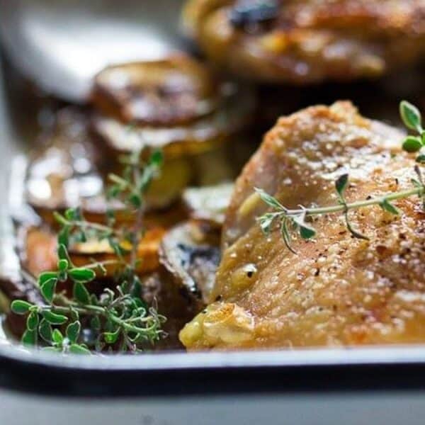 Roasted chicken thighs in a baking dish, topped with sprigs of fresh thyme on a layer of sliced vegetables.