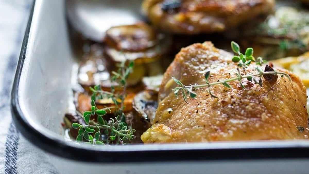 Roasted chicken thighs in a baking dish, topped with sprigs of fresh thyme on a layer of sliced vegetables.
