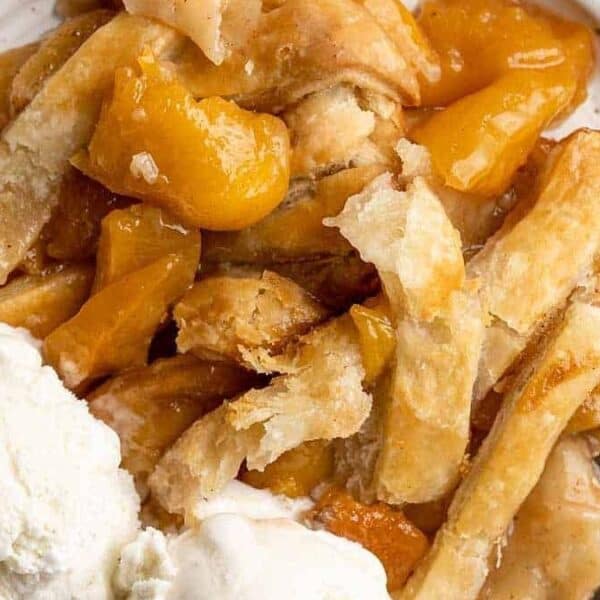 Peach cobbler with a flaky crust on a plate, two scoops of vanilla ice cream beside it, and a spoon.