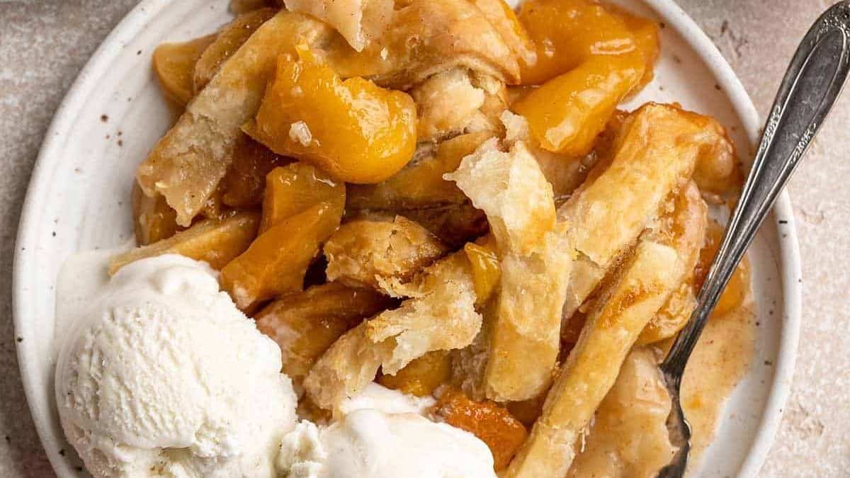 Peach cobbler with a flaky crust on a plate, two scoops of vanilla ice cream beside it, and a spoon.