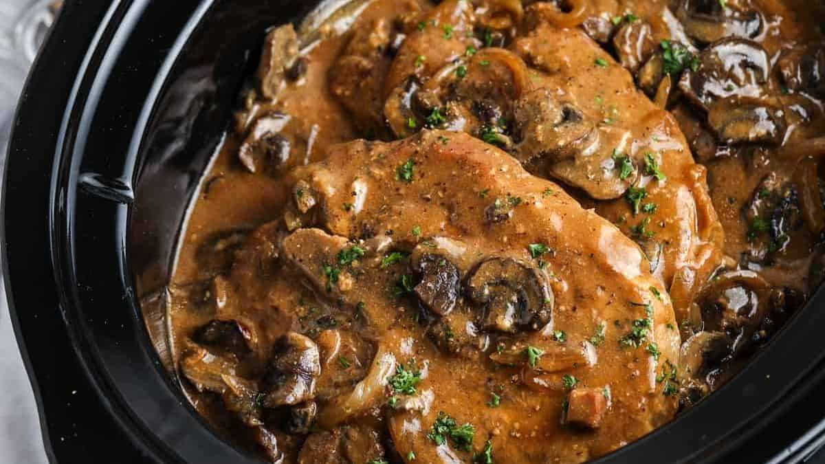 Sliced beef topped with creamy brown mushroom sauce and chopped herbs in a black slow cooker.
