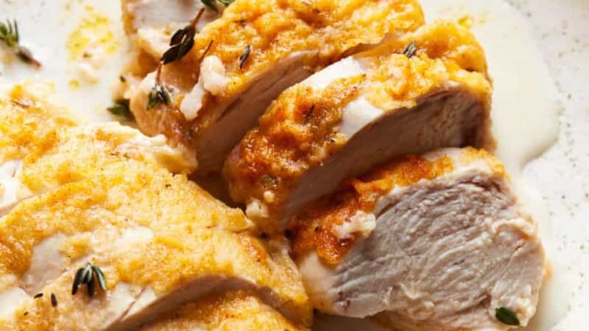 Sliced baked chicken breast with a golden-brown cheese and herb crust, alongside a creamy sauce.