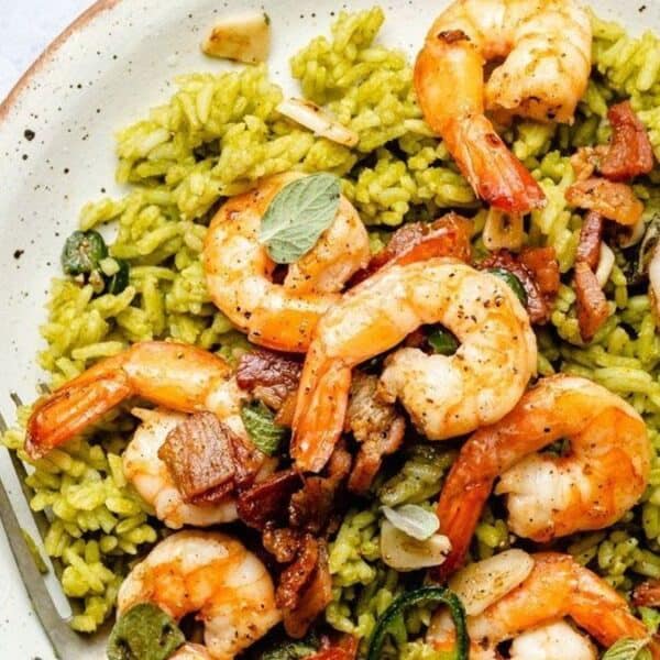 A plate of yellow-green rice topped with cooked shrimp, bacon pieces, fresh herbs, and a lime wedge.