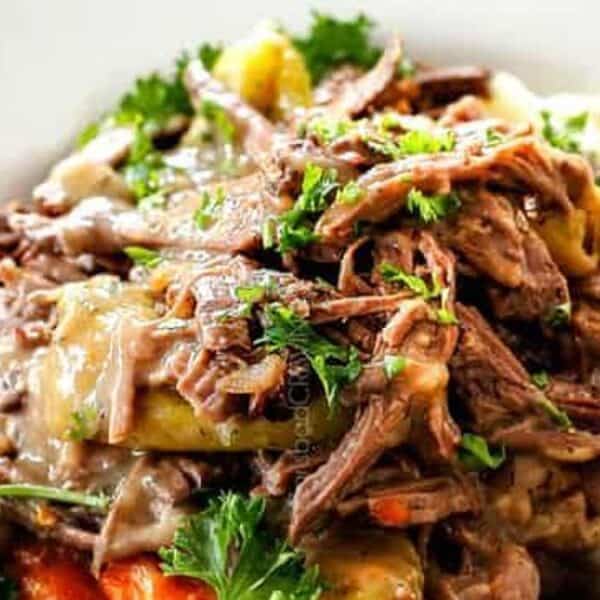 Shredded pot roast topped with gravy, carrots, peppers, and parsley on a bed of mashed potatoes, all on a white plate.