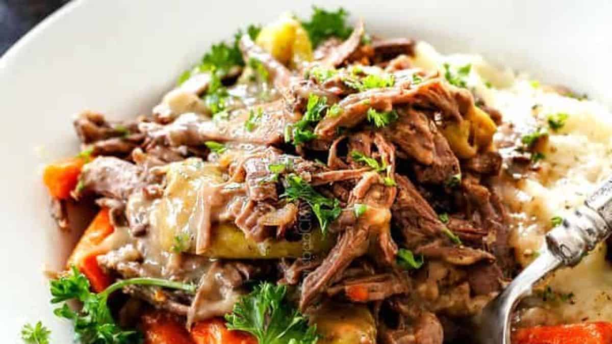 Shredded pot roast topped with gravy, carrots, peppers, and parsley on a bed of mashed potatoes, all on a white plate.