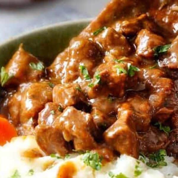 A bowl of beef stew with carrots over mashed potatoes, garnished with chopped parsley, being spooned with a wooden spoon.