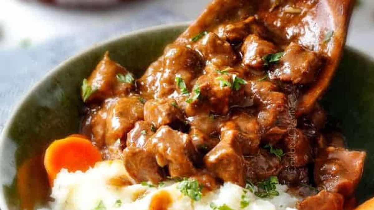 A bowl of beef stew with carrots over mashed potatoes, garnished with chopped parsley, being spooned with a wooden spoon.