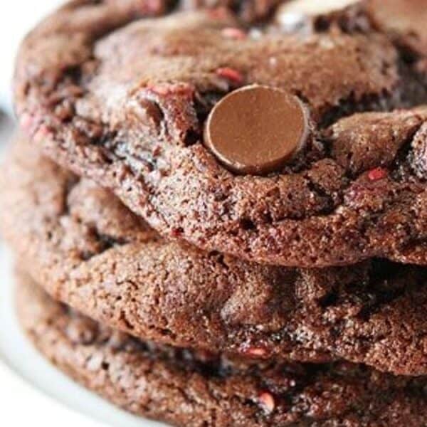 Stack of three chocolate chip cookies on a white plate, with some chocolate chips blurred in the background.
