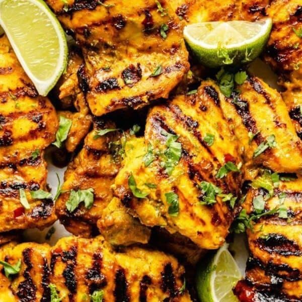 Grilled chicken pieces with char marks, topped with chopped herbs and lime wedges, arranged on a white plate.