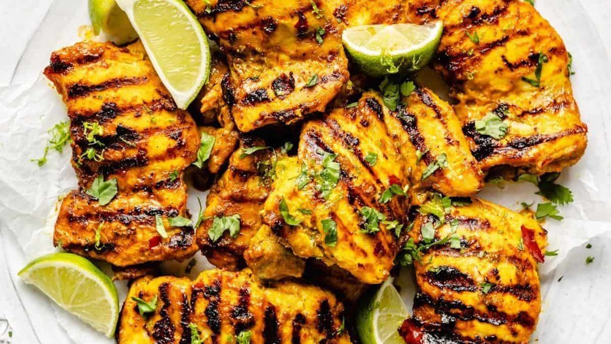 Grilled chicken pieces with char marks, topped with chopped herbs and lime wedges, arranged on a white plate.