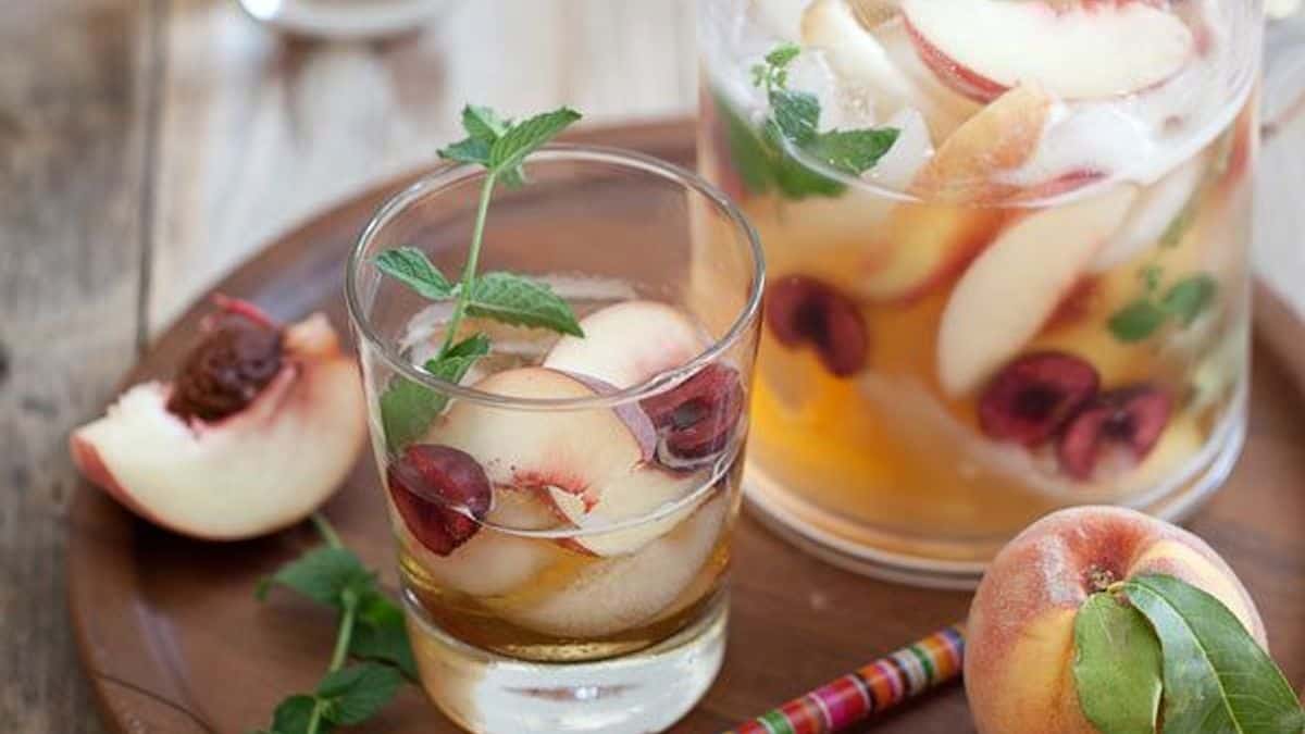 A glass and pitcher filled with iced fruit drink, sliced peaches, cherries, mint leaves, ice, and fresh fruit on a wooden tray.