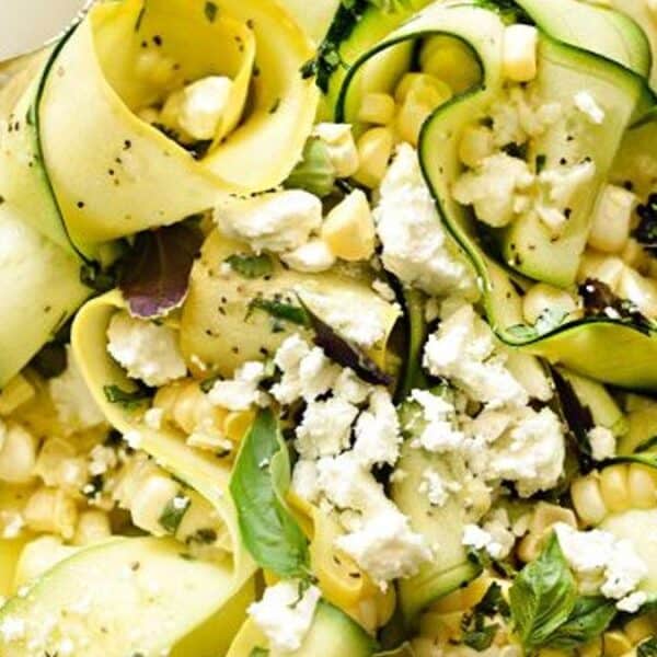 A plate containing zucchini ribbons, corn, crumbled feta cheese, fresh herbs, and a fork placed beside the salad.