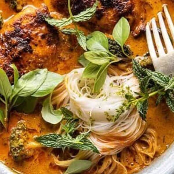 A bowl of red curry with grilled chicken, noodles, basil, mint, and broccoli, with a fork and spoon beside it.