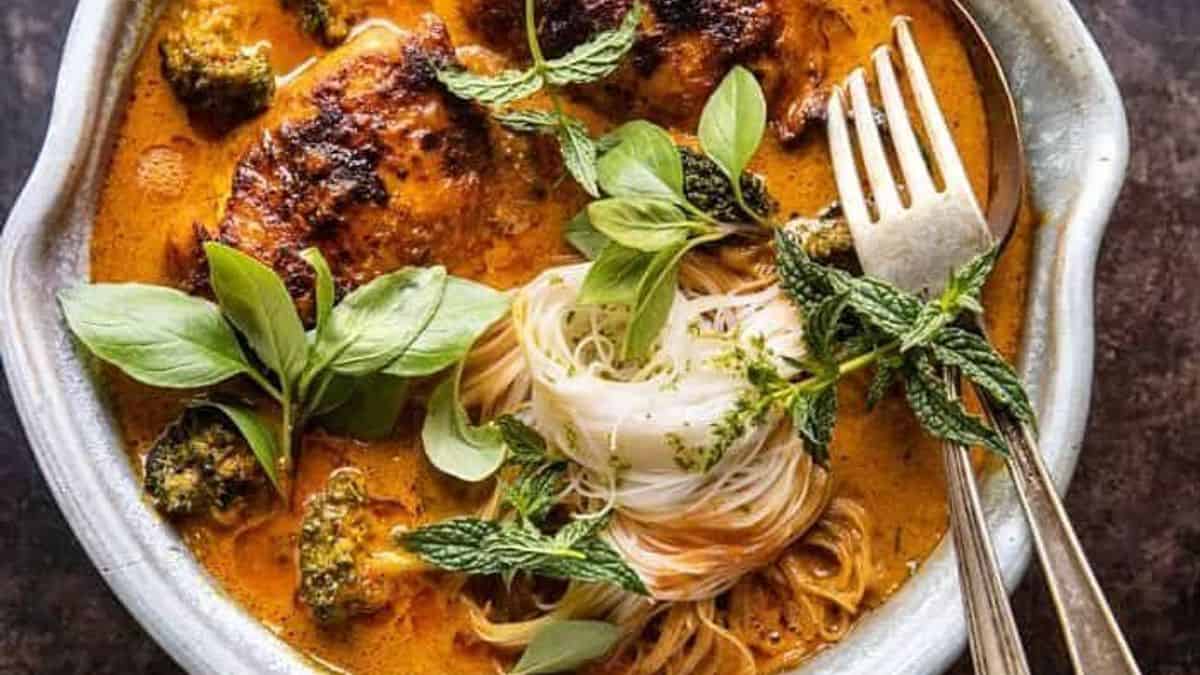 A bowl of red curry with grilled chicken, noodles, basil, mint, and broccoli, with a fork and spoon beside it.