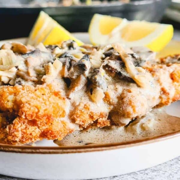 Breaded chicken cutlet with creamy mushroom sauce, two lemon wedges, served on a white plate.