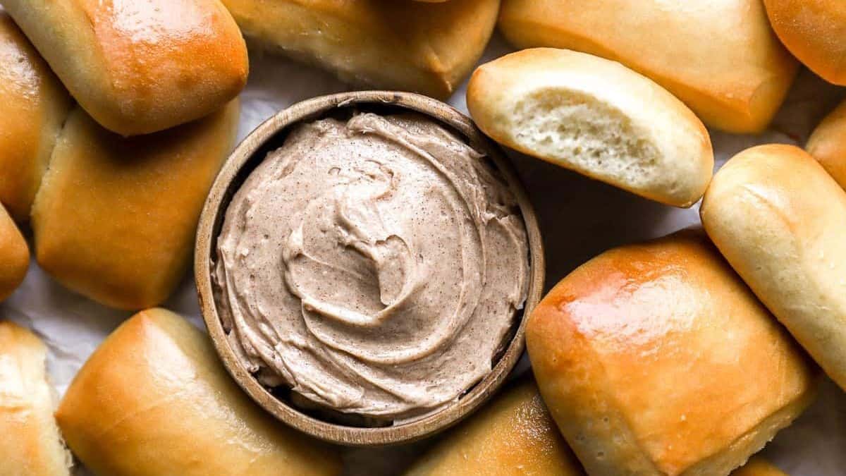 A bowl of cinnamon butter surrounded by several golden brown dinner rolls.