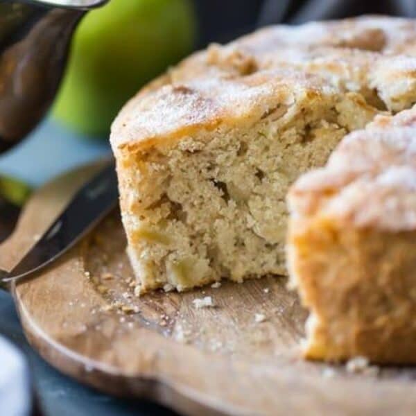 A round apple cake with a slice removed sits on a wooden board, with a knife nearby and green apples in the background.