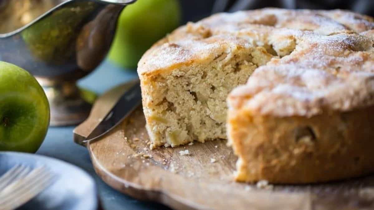 A round apple cake with a slice removed sits on a wooden board, with a knife nearby and green apples in the background.