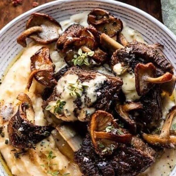 A plate of mashed potatoes with roasted meat, sautéed mushrooms, fresh herbs, and creamy sauce on a wooden table.