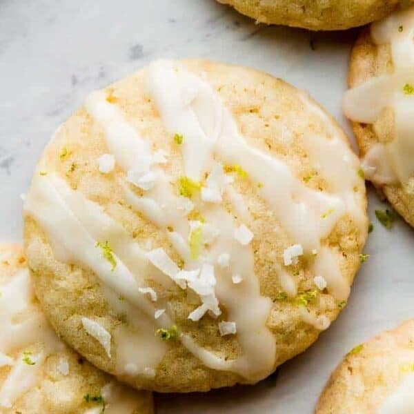 Round cookies with white icing drizzle, shredded coconut, and lime zest on a light-colored surface.