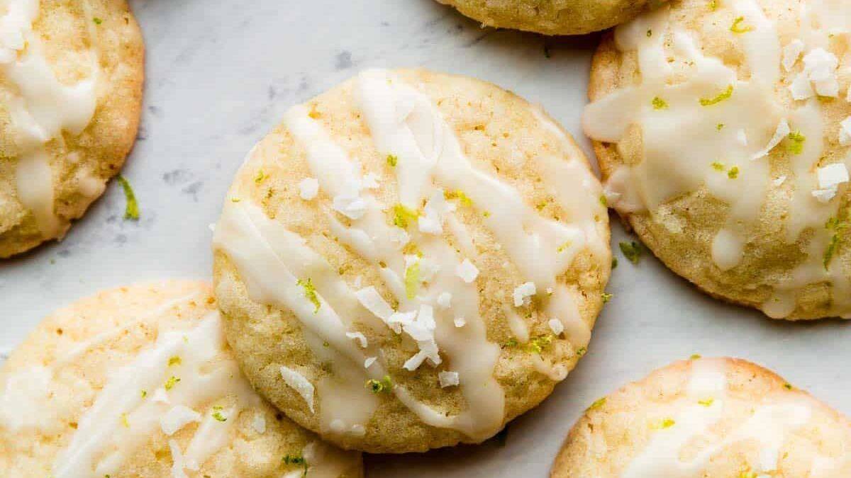 Round cookies with white icing drizzle, shredded coconut, and lime zest on a light-colored surface.