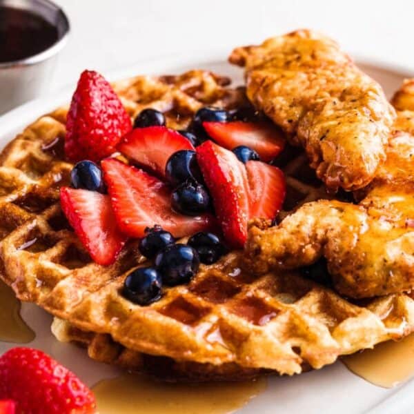 Waffle with fried chicken, sliced strawberries, blueberries, and syrup on a plate, plus a cup of syrup beside it.