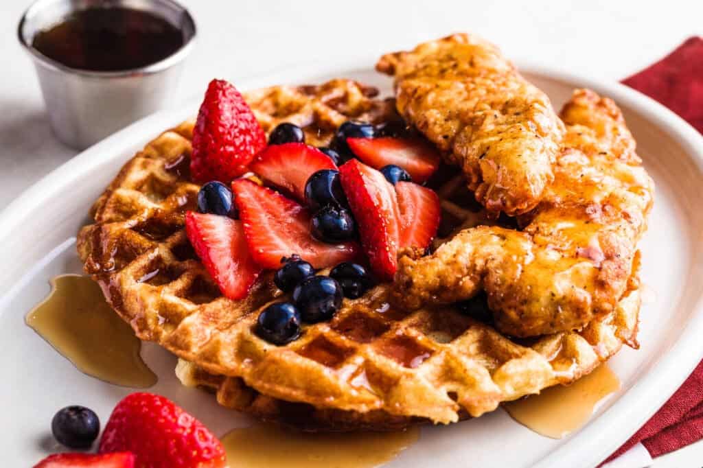 Waffle with fried chicken, sliced strawberries, blueberries, and syrup on a plate, plus a cup of syrup beside it.