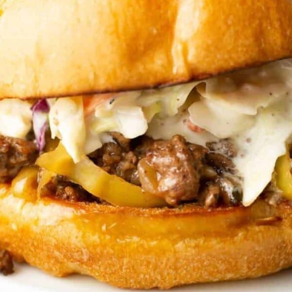 A sandwich with ground beef, yellow peppers, and coleslaw on a toasted bun sits on a white plate.