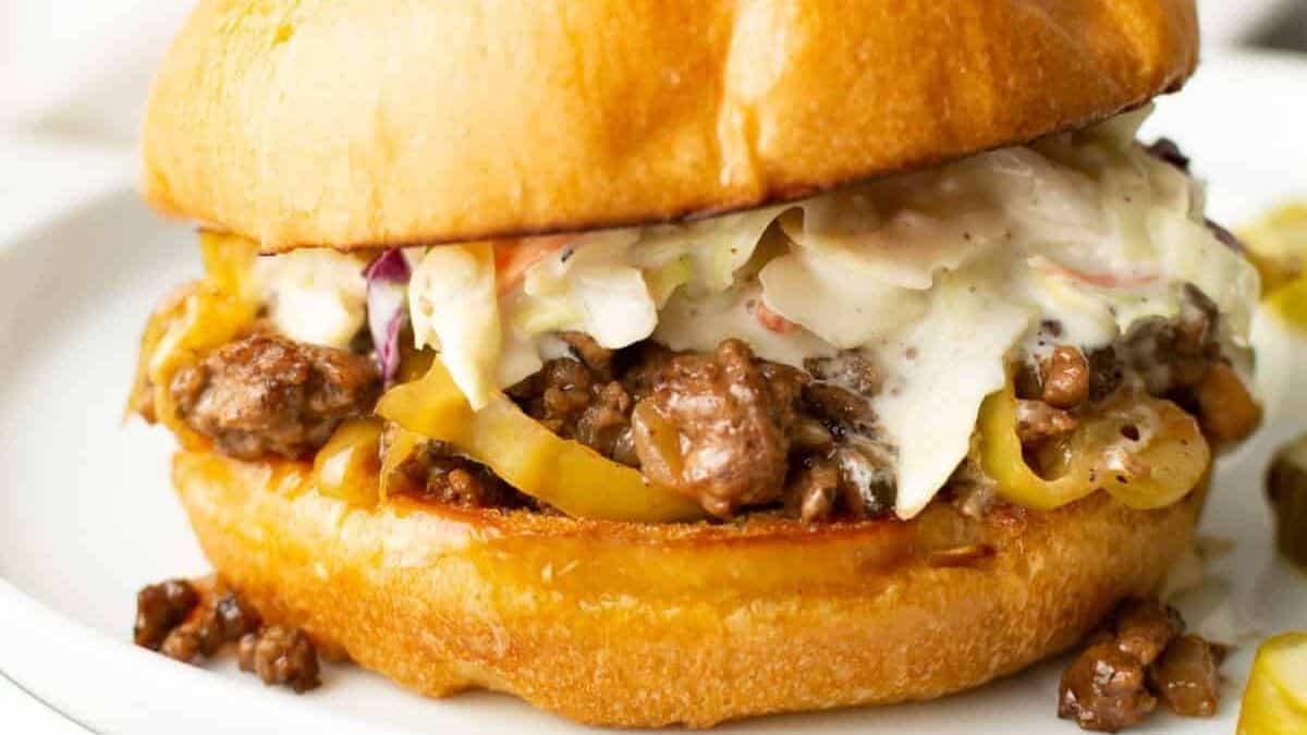 A sandwich with ground beef, yellow peppers, and coleslaw on a toasted bun sits on a white plate.