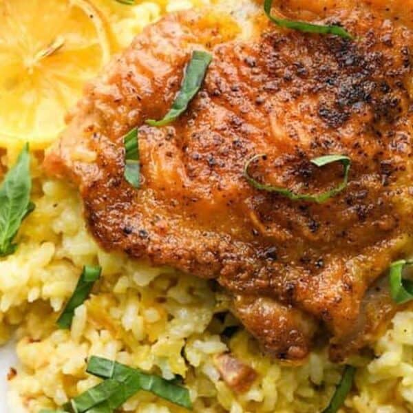 Plate with roasted chicken thigh on yellow rice, garnished with lemon slices and fresh basil, fork placed beside the plate.