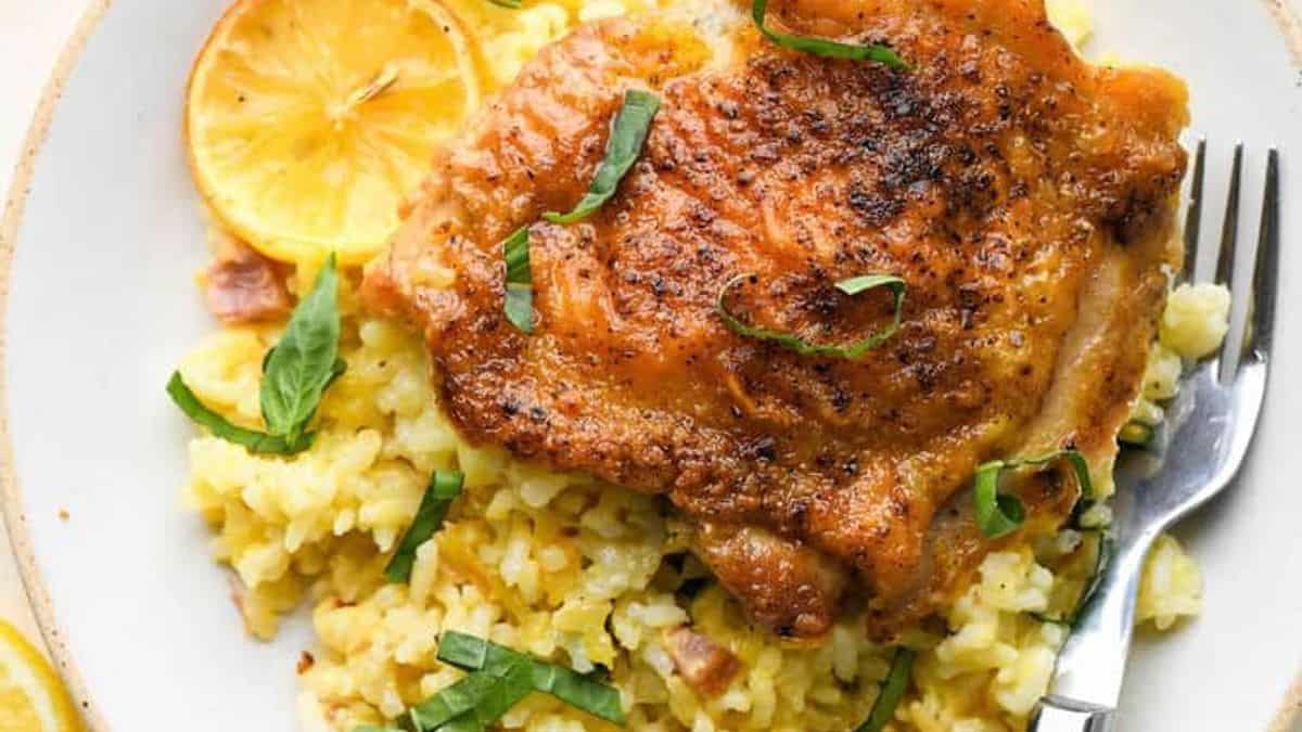 Plate with roasted chicken thigh on yellow rice, garnished with lemon slices and fresh basil, fork placed beside the plate.