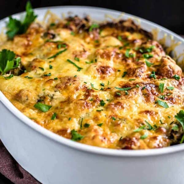 A white oval dish holds a casserole topped with melted cheese and parsley, set on a dark surface.