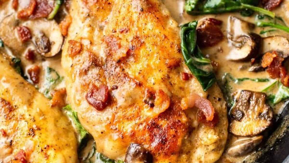 Chicken breasts, creamy sauce, mushrooms, spinach, and bacon pieces cooked together in a skillet.