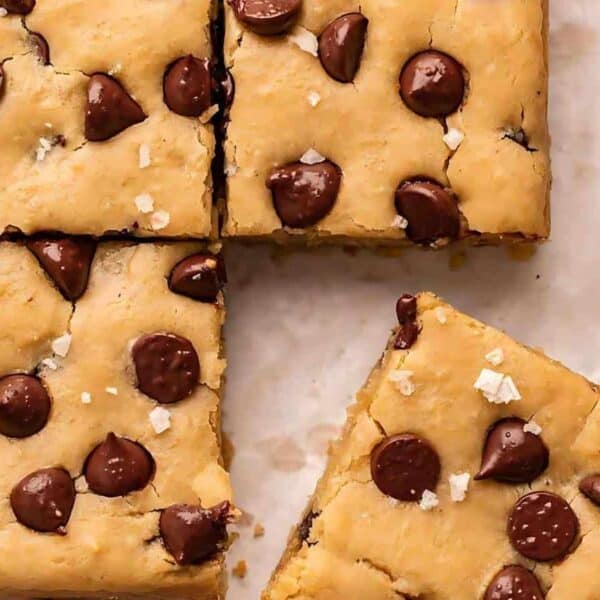 Squares of blondie bars with chocolate chips on parchment paper, one bar set apart and a few chocolate chips scattered nearby.