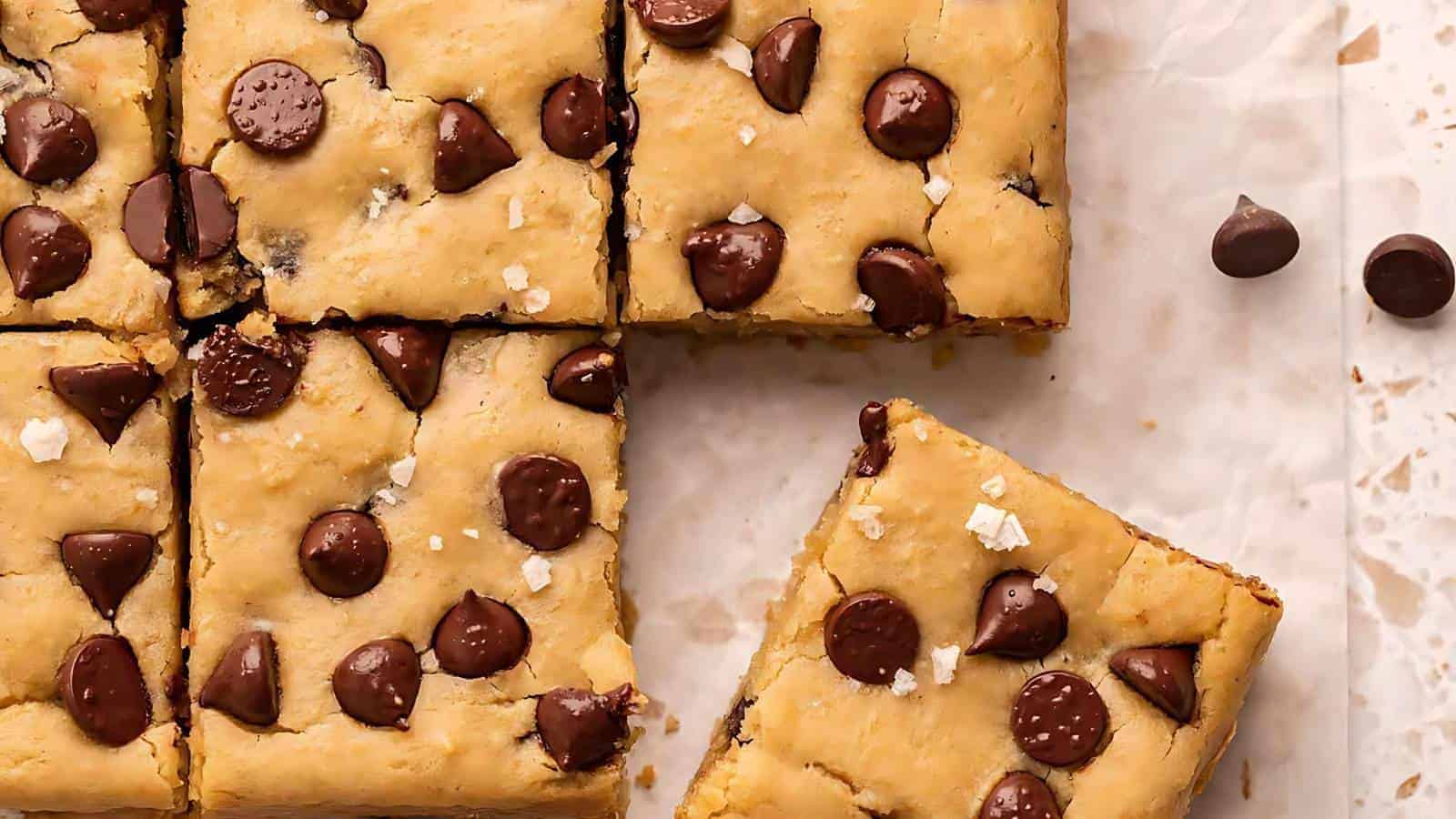 Squares of blondie bars with chocolate chips on parchment paper, one bar set apart and a few chocolate chips scattered nearby.