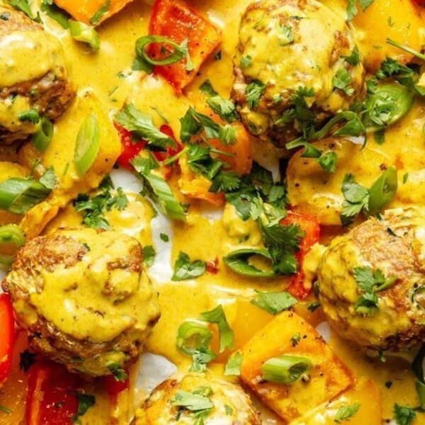 Meatballs topped with herbs and green onions in yellow sauce, diced red and yellow bell peppers, and a spoon with sauce on the side.
